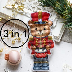 Könnte beinhalten: Ein Kreuzstich-Weihnachtsschmuck in Form eines Teddybären, der als Spielzeugsoldat gekleidet ist und einen roten Hut und eine Jacke trägt. Der Text "3 in 1" befindet sich in einem schwarzen Kreis. Weitere Bastelmaterialien im Hintergrund.