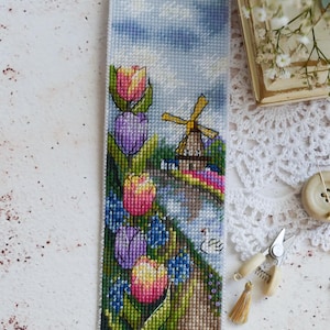 Könnte beinhalten: Ein rechteckiges Lesezeichen in Kreuzstichoptik mit einer Landschaftsszene, die eine gelbe Windmühle, einen blauen Himmel mit weißen Wolken sowie pinke, gelbe und violette Tulpen und blaue Blumen auf einem grünen Pfad zeigt.