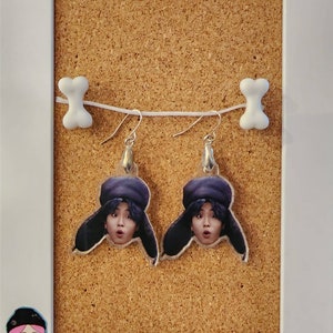 May include: A pair of silver dangle earrings with clear acrylic charms featuring a person's face wearing a black and purple hat. The earrings are hanging from a cork board with the text "Step Made Something" at the bottom.