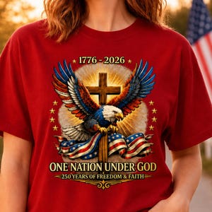 May include: A red short-sleeve t-shirt featuring a graphic of a bald eagle with spread wings perched on a wooden cross, draped with an American flag. The text reads 1776-2026, ONE NATION UNDER GOD, and 250 YEARS OF FREEDOM & FAITH.
