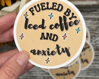 May include: A round sticker with a beige background and black text that reads "Fueled by Iced Coffee and Anxiety". The sticker has small white stars around the text.