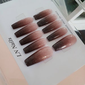 Op de afbeelding: Set van tien lange, amandelvormige press-on nagels met een verloopdesign. De nagels zijn lichtbruin aan de punt en vervagen naar donkerbruin aan de basis. De nagels zijn versierd met glitter. De tekst "HANDMADE PRESS ON NAILS" en "LNN" staat op de kaart gedrukt.