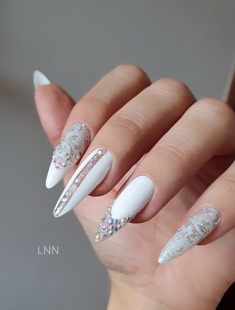 Op de afbeelding: Een hand met lange, witte, stiletto-vormige nagels. Sommige nagels zijn versierd met zilveren en heldere kristallen details. E&eacute;n nagel heeft een zilveren streep. De nagels hebben een glanzende afwerking en zijn versierd met een bloemmotief.