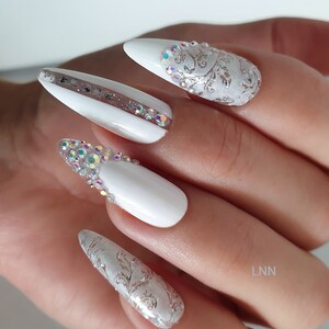 Op de afbeelding: Close-up van een hand met witte, amandelvormige acrylnagels. Sommige nagels hebben zilveren bloemmotieven en iriserende strass-steentjes. E&eacute;n nagel heeft een streep glitter en strass-steentjes. De nagels zijn professioneel gedaan.