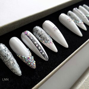 Op de afbeelding: Set witte, lange, puntige valse nagels. Sommige nagels hebben zilveren glitter, strass en zwarte accenten. De nagels zijn gerangschikt in een zwarte doos met een zilverkleurige rand. De initialen "LNN" zijn linksonder zichtbaar.