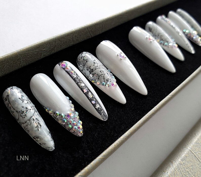 Op de afbeelding: Set witte, lange, puntige valse nagels. Sommige nagels hebben zilveren glitter, strass en zwarte accenten. De nagels zijn gerangschikt in een zwarte doos met een zilverkleurige rand. De initialen "LNN" zijn linksonder zichtbaar.