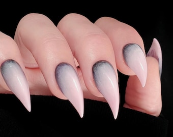 May include: Close-up of a hand with long, pointed, artificial nails. The nails have a gradient design, transitioning from a dark gray at the base to a soft pink at the tips. The background is solid black.