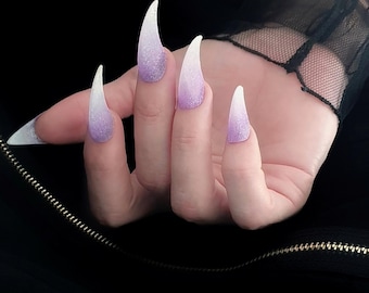 May include: Long, pointed artificial nails with a gradient design. The nails transition from a white tip to a shimmering lavender base. The hand is set against a dark background, highlighting the nail art.