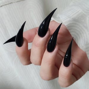 Puede incluir: Primer plano de una mano con uñas stiletto largas y brillantes de color negro. Las uñas son puntiagudas y reflejan la luz, creando una apariencia elegante y dramática. Fondo blanco.
