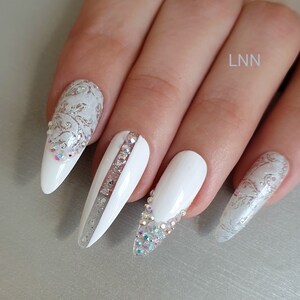Op de afbeelding: Witte, amandelvormige kunstnagels met ingewikkelde ontwerpen. Sommige nagels hebben zilveren glitteraccenten en strass-steentjes. E&eacute;n nagel heeft een verticale streep van strass-steentjes. De nagels zijn versierd met een bloemmotief.