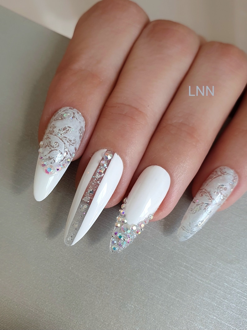 Op de afbeelding: Witte, amandelvormige kunstnagels met ingewikkelde ontwerpen. Sommige nagels hebben zilveren glitteraccenten en strass-steentjes. E&eacute;n nagel heeft een verticale streep van strass-steentjes. De nagels zijn versierd met een bloemmotief.