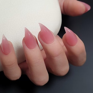 Puede incluir: Primer plano de una mano con uñas acrílicas largas, puntiagudas y rosas. Las uñas tienen un acabado brillante y un aspecto translúcido. Los dedos están ligeramente curvados, mostrando la forma y el color de las uñas.