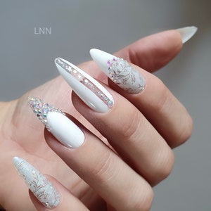 Op de afbeelding: Witte, amandelvormige kunstnagels met ingewikkelde ontwerpen. Sommige nagels hebben een zilveren streep, andere zijn versierd met iriserende strass-steentjes en bloemmotieven. De nagels zijn lang en elegant, perfect voor een speciale gelegenheid.