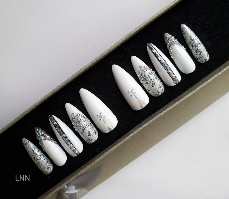 Op de afbeelding: Een set witte, lange, puntige valse nagels met verschillende ontwerpen. Sommige nagels hebben zilveren glitter, strass en ingewikkelde bloempatronen. De nagels zijn gerangschikt in een doos.