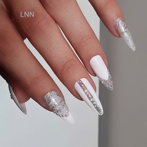 Op de afbeelding: Witte stiletto nagels met zilveren en heldere kristallen accenten. Sommige nagels hebben een zilveren bloemenpatroon. E&eacute;n nagel heeft een zilveren streep met heldere kristallen. De letters "LNN" zijn zichtbaar.