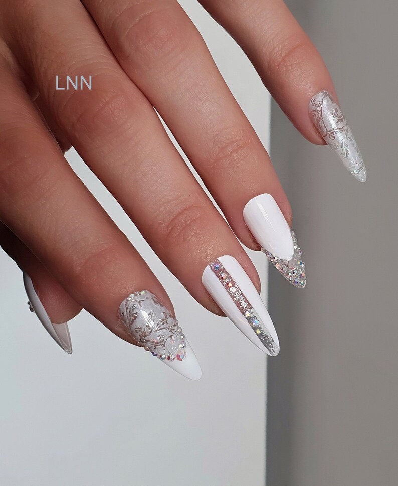 Op de afbeelding: Witte stiletto nagels met zilveren en heldere kristallen accenten. Sommige nagels hebben een zilveren bloemenpatroon. E&eacute;n nagel heeft een zilveren streep met heldere kristallen. De letters "LNN" zijn zichtbaar.