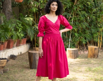 May include: A woman wearing a pink wrap dress with a gathered skirt and puffed sleeves. She is standing in a garden setting.