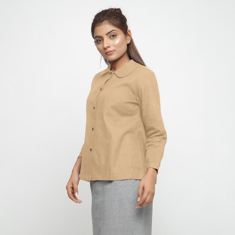 May include: A light brown collared shirt with long sleeves and a button-up front. The shirt has a peter pan collar.