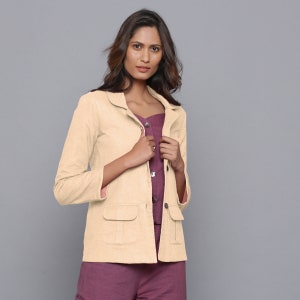 May include: A light beige blazer with two front pockets and a notched collar. The blazer is worn over a plum-colored top and pants. The sleeves are three-quarter length. The background is a solid gray.