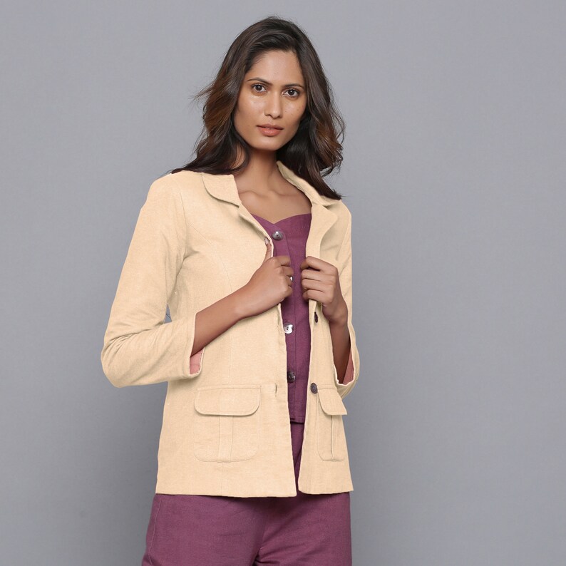 May include: A light beige blazer with two front pockets and a notched collar. The blazer is worn over a plum-colored top and pants. The sleeves are three-quarter length. The background is a solid gray.