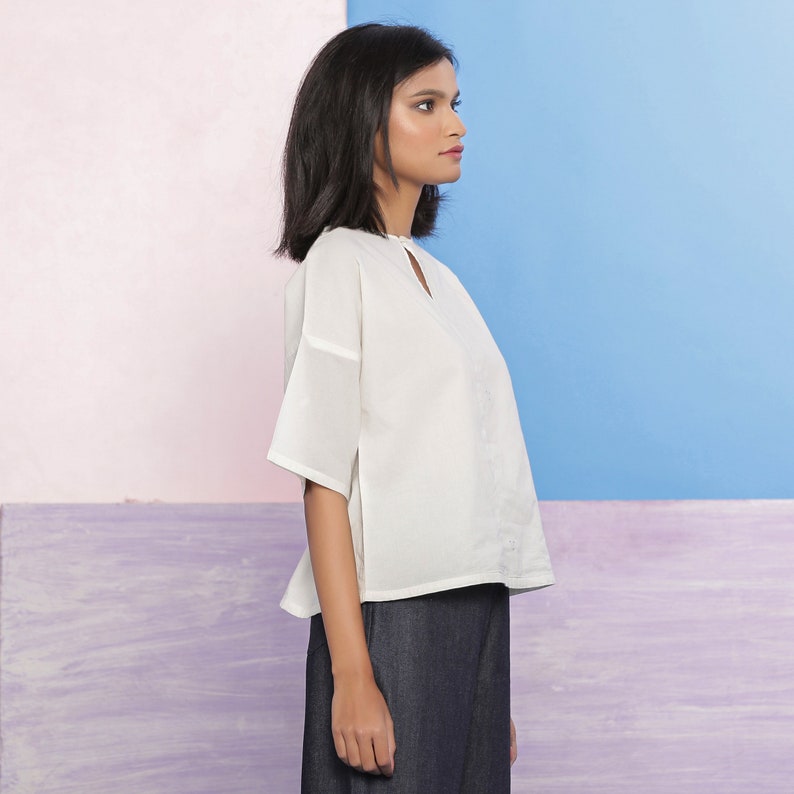 May include: A white, short-sleeved, loose-fitting blouse with a keyhole neckline. The blouse is worn with dark blue pants.