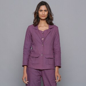 May include: A woman wearing a matching purple blazer, top, and pants set. The blazer has two front pockets and button closures. The outfit is a solid color and appears to be made of a linen or cotton blend. The background is a solid gray.