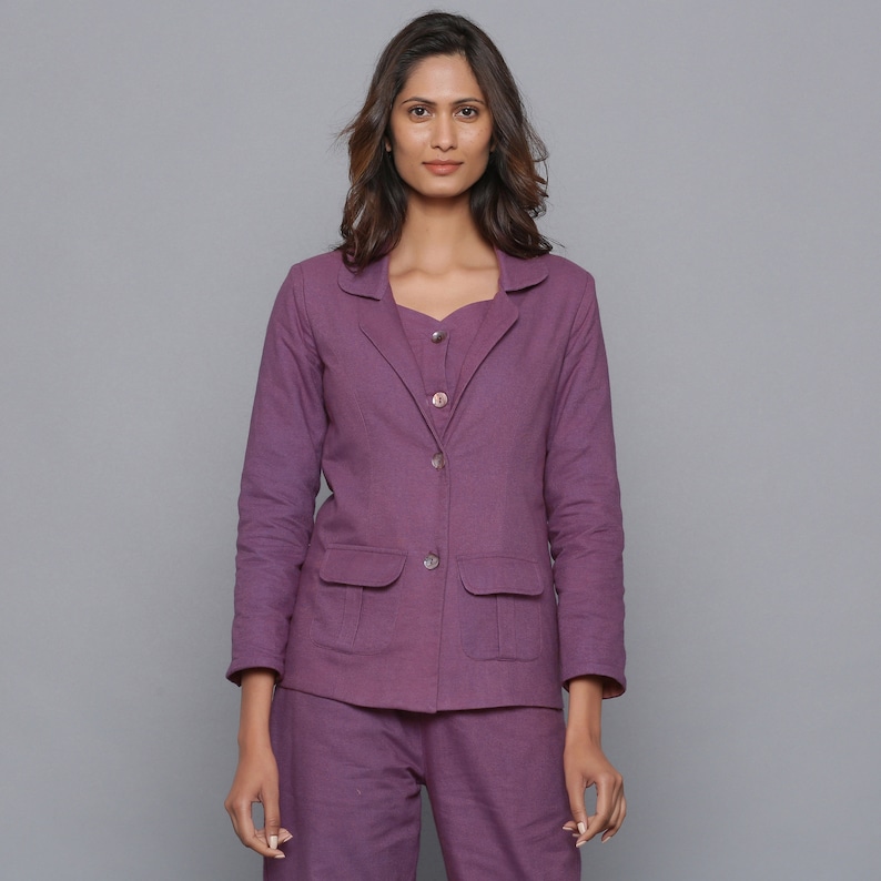 May include: A woman wearing a matching purple blazer, top, and pants set. The blazer has two front pockets and button closures. The outfit is a solid color and appears to be made of a linen or cotton blend. The background is a solid gray.