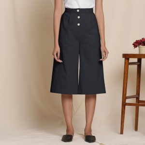 May include: Black culottes with a high waist and three large, round, cream-colored buttons. The wide-leg pants are knee-length and made of a solid, dark fabric. The model is wearing black flats.