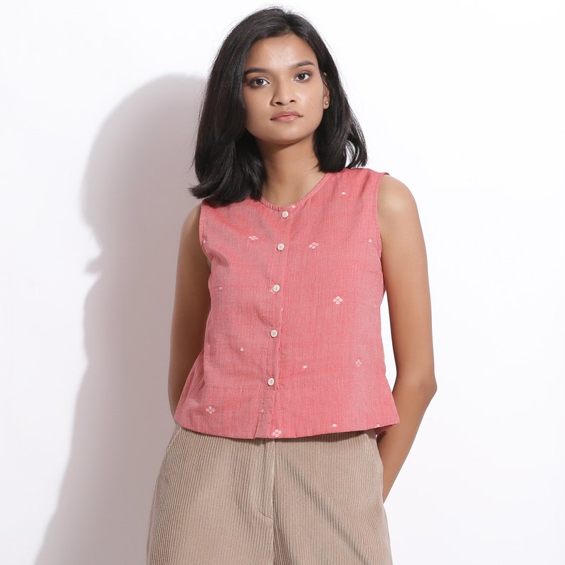 Puede incluir: Una blusa rosa sin mangas con botones y un estampado floral blanco. La blusa est&aacute; hecha de una tela ligera y tiene un corte holgado.