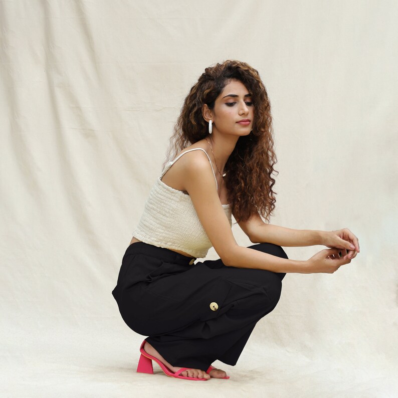 May include: A woman wearing a white crop top and black wide-leg pants with a gold belt buckle. She is wearing pink heeled sandals and has long curly brown hair.