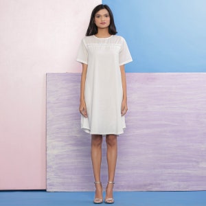 Puede incluir: Un vestido blanco de manga corta con un ribete de encaje alrededor del escote. El vestido tiene un corte holgado y llega hasta las rodillas. La modelo lleva tacones plateados.