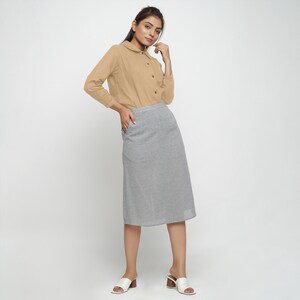 May include: A woman is wearing a light brown long-sleeved collared shirt with a gray midi skirt. She is standing with her hand in her pocket and looking at the camera.