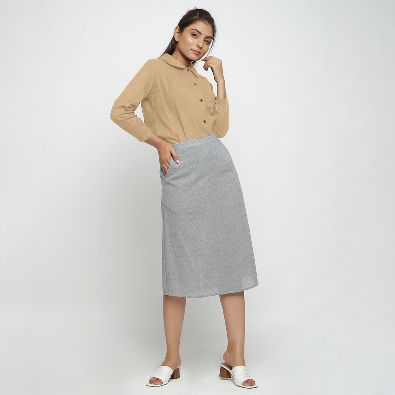 May include: A woman is wearing a light brown long-sleeved collared shirt with a gray midi skirt. She is standing with her hand in her pocket and looking at the camera.