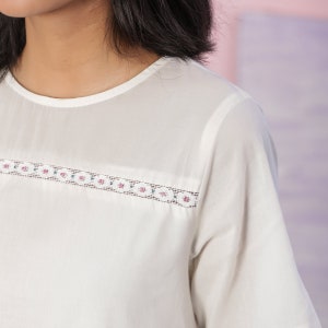 Puede incluir: Una blusa blanca de manga corta con un delicado ribete de encaje alrededor del escote. El ribete de encaje es de color rosa claro y verde.