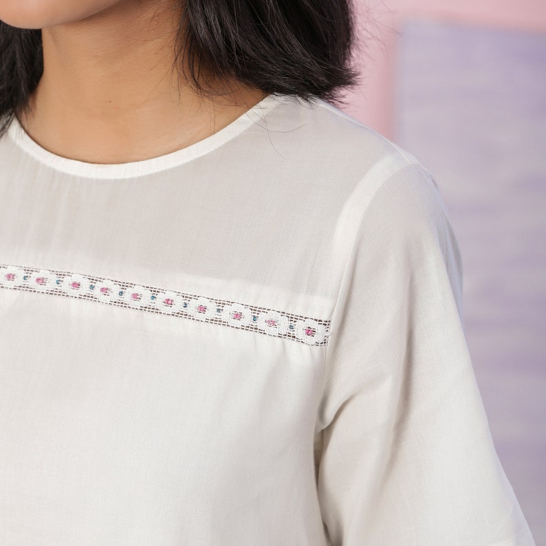 Puede incluir: Una blusa blanca de manga corta con un delicado ribete de encaje alrededor del escote. El ribete de encaje es de color rosa claro y verde.