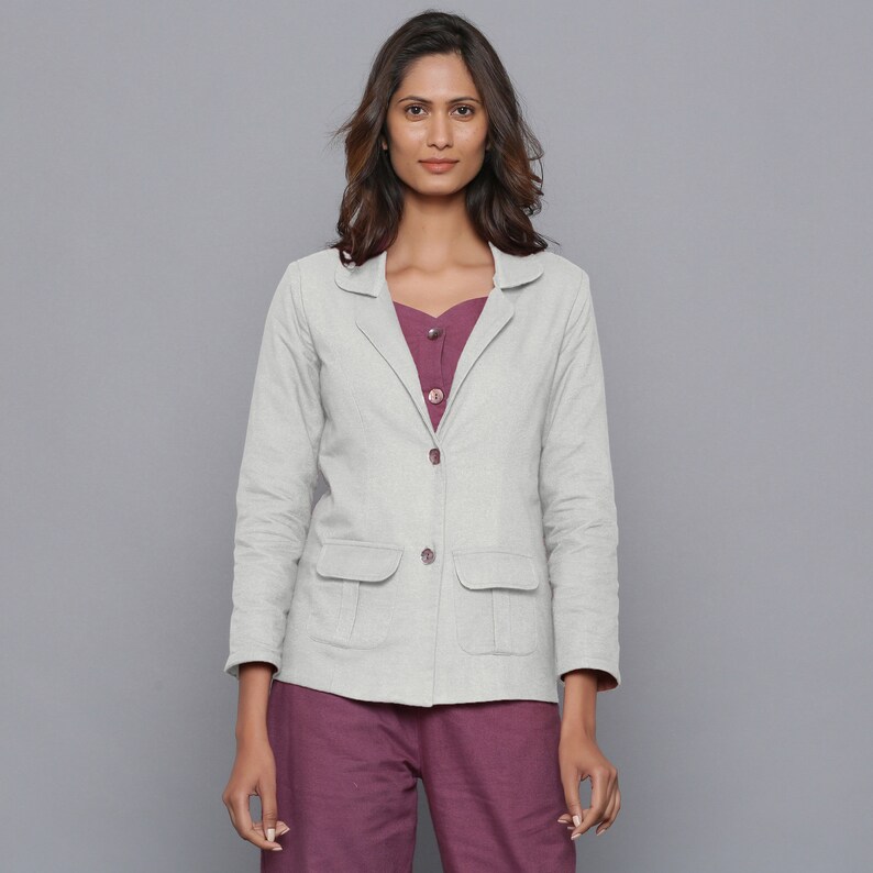 May include: A light gray blazer with two front pockets and matching buttons. The blazer is worn over a plum-colored top and paired with plum-colored pants. The outfit is set against a gray background.