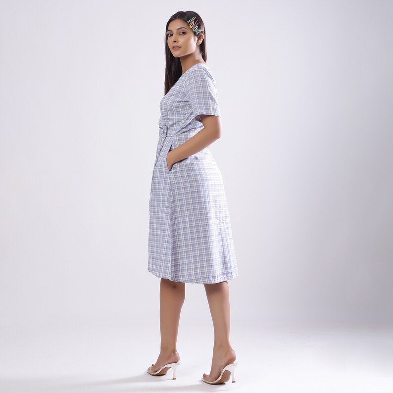 May include: A woman wearing a light blue and white plaid dress with a short sleeve and a belt. She is standing with her hands in her pockets and looking over her shoulder.