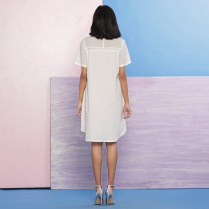Puede incluir: Un vestido blanco de manga corta con un ribete de encaje alrededor del escote. El vestido es de largo hasta la rodilla y tiene un corte holgado. La modelo lleva el vestido con tacones plateados.