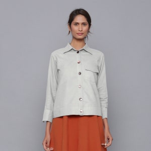 Puede incluir: Una camisa de manga larga de color gris claro con botones y un bolsillo único en el pecho. La camisa tiene un corte holgado y está hecha de una tela suave y ligera.