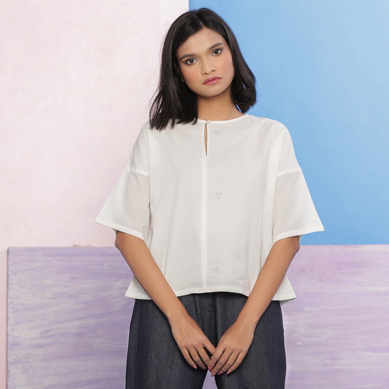 May include: A white cotton blouse with a keyhole neckline and three small embroidered details. The blouse has a relaxed fit and short sleeves.