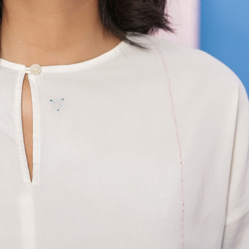 May include: White button-down shirt with a small keyhole opening at the neckline and a thin pink stitching detail along the shoulder seam.