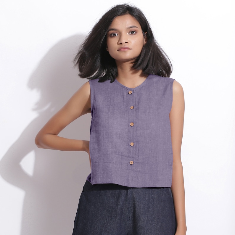 Puede incluir: Una blusa de lino morada sin mangas con cierre de botones en la parte delantera. La blusa tiene un escote redondo y un dobladillo recortado.