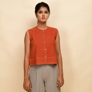 Puede incluir: Una blusa sin mangas de color &oacute;xido con botones y cuello redondo. La blusa tiene una longitud recortada y un dobladillo ligeramente acampanado. La modelo lleva la blusa con pantalones grises.