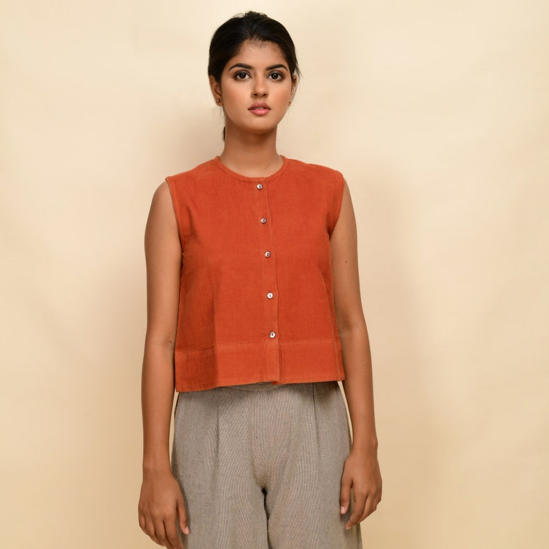 Puede incluir: Una blusa sin mangas de color &oacute;xido con botones y cuello redondo. La blusa tiene una longitud recortada y un dobladillo ligeramente acampanado. La modelo lleva la blusa con pantalones grises.