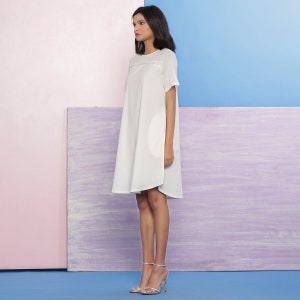 Puede incluir: Un vestido blanco, de manga corta, de corte A, con un ribete de encaje alrededor del escote. El vestido lo lleva una persona de pie sobre una superficie azul.