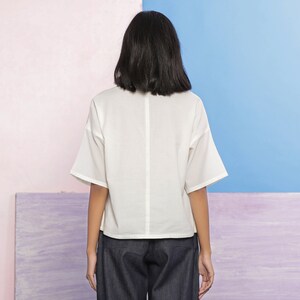 May include: A white, short-sleeved, collared blouse with a relaxed fit. The blouse has a slightly cropped hemline and is made of a lightweight fabric.