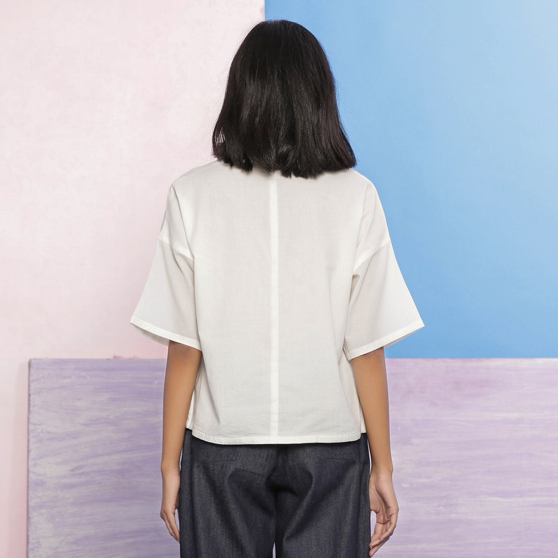 May include: A white, short-sleeved, collared blouse with a relaxed fit. The blouse has a slightly cropped hemline and is made of a lightweight fabric.