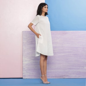 Puede incluir: Un vestido blanco, de manga corta, de corte A, con un ribete de encaje alrededor del escote. El vestido se muestra en una modelo con un fondo rosa claro a la izquierda y un fondo morado claro a la derecha.