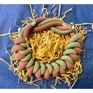 Può includere: Una collana spessa e intrecciata realizzata in due tonalità di filato, rosa e verde. La collana è realizzata in un materiale morbido e testurizzato e ha una chiusura argentata.