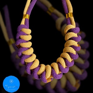 Può includere: Una collana annodata di colore viola e beige con accenti dorati. La collana è realizzata con due fili di cordoncino intrecciati tra loro.